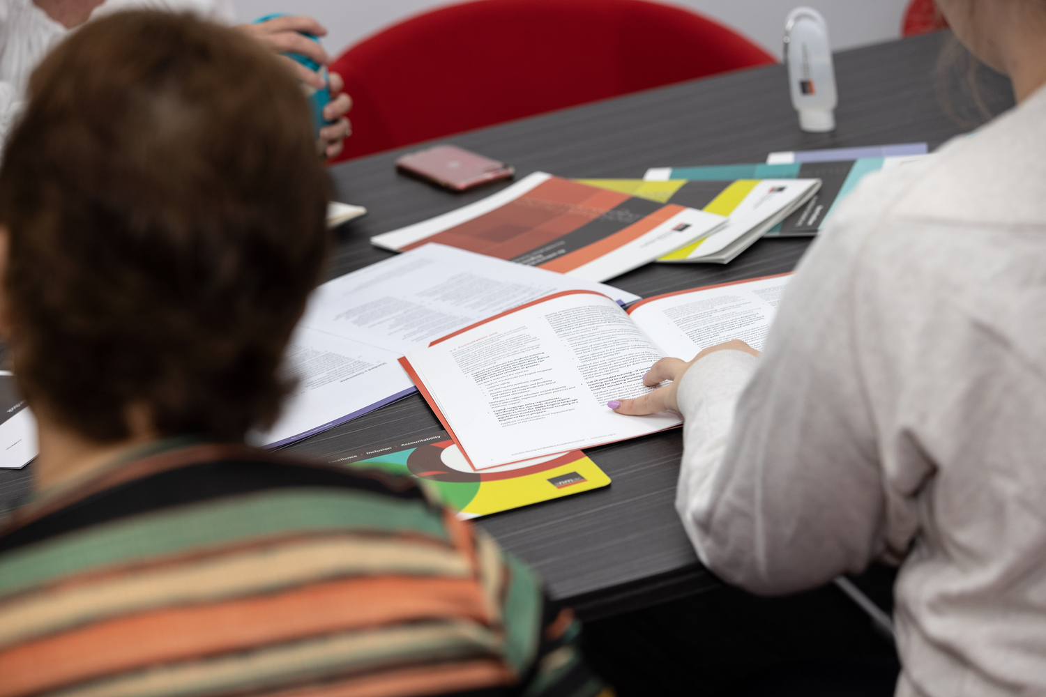 Two people sit at a table reviewing printed documents and booklets. One person is reading an open booklet while various colourful reports and papers are spread across the dark tabletop.